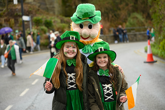 2 young girls dressed in green outfits posing with a person dressed as a leprechaun outdoors on a road.
