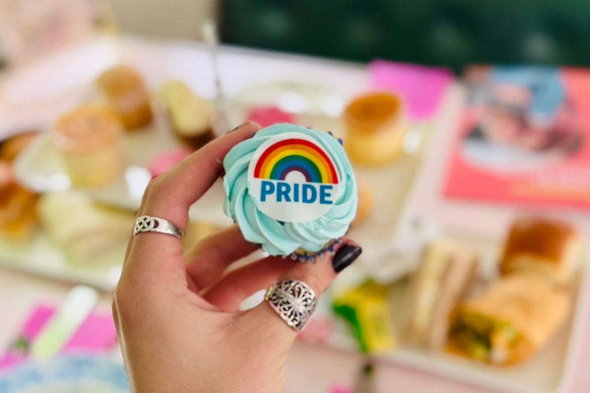 Pride Month - Afternoon Tea Trip - a hand holding out a small cup cake with swirl of icing on and a small rainbow logo, against blurry background of tea setting