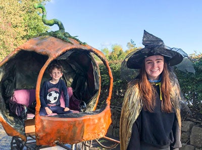 A smiling girl in black witches hat and costume with orange pumpkin style small carriage behind with young boy seated in it.