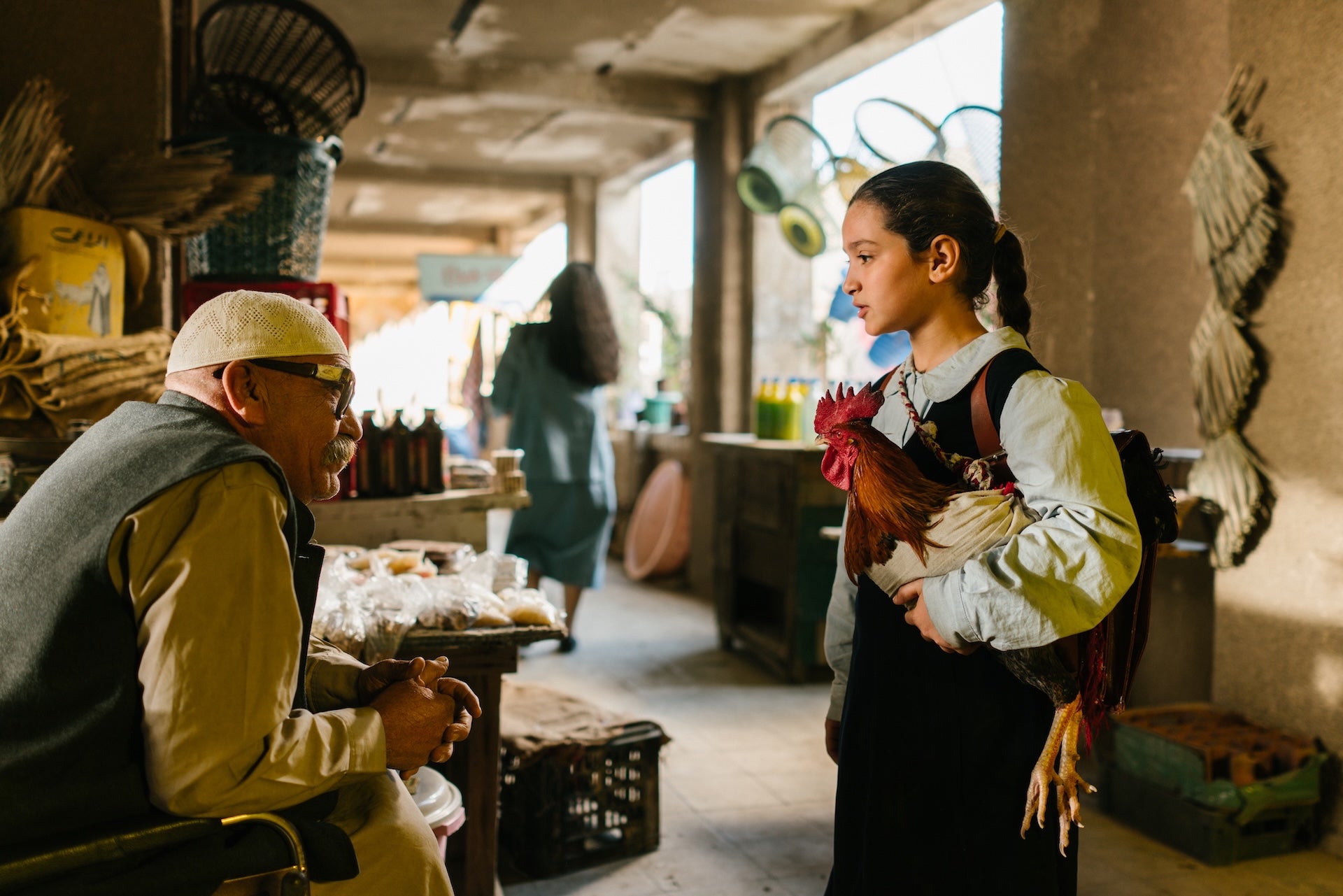 A girl with a chicken is talking a man at a market stall.