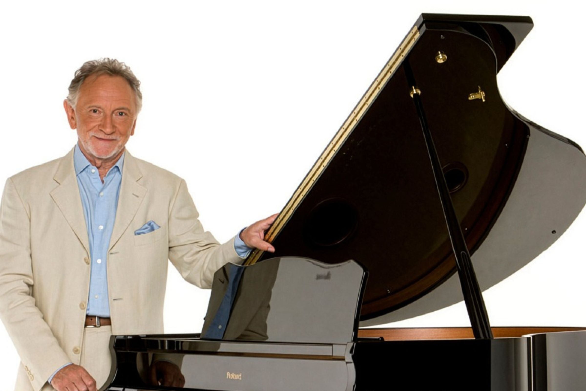Phil Coulter in Concert Dec 2026, a smiling man in light coloured suit standing beside a grand piano with the lid open.
