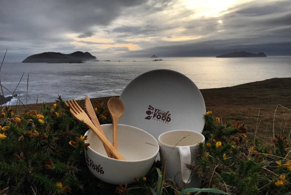 A white bowl, plate and mug holding set of wooden cutlery set against sea and rocky background.