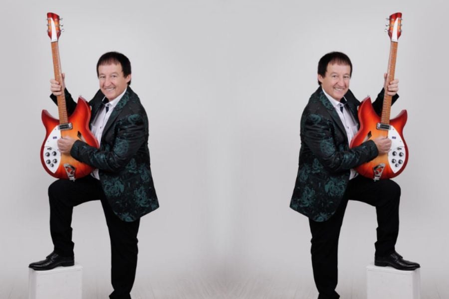 A smiling man in dark suit with one foot up on small white block, holding up a guitar vertically 