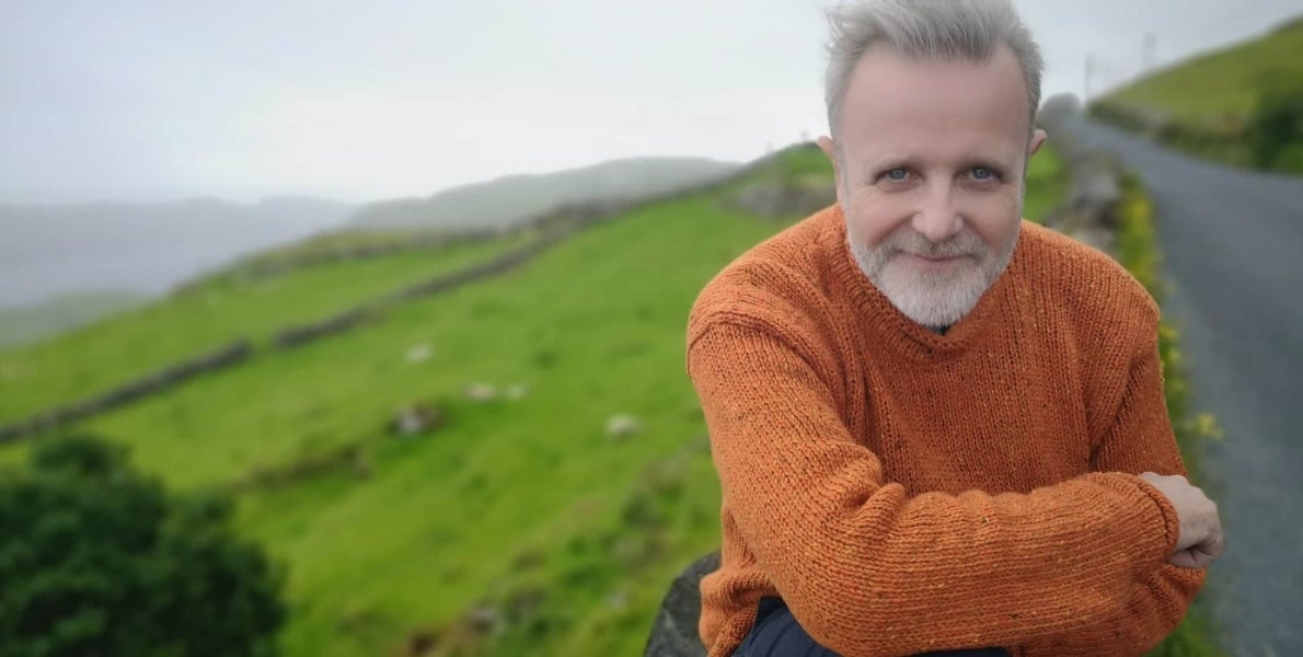 A smiling man with crossed arms outdoors with background of green hills