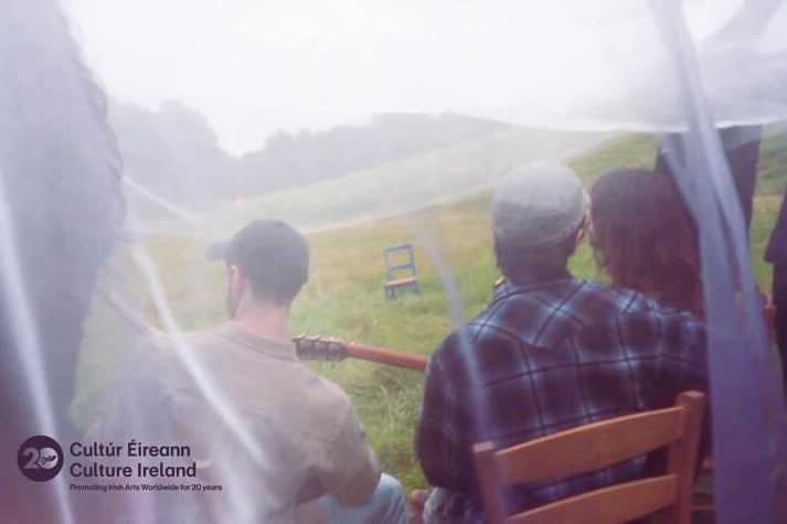 rear view of 3 people seated on wooden chairs in a grassy field, viewed through white net curtain.