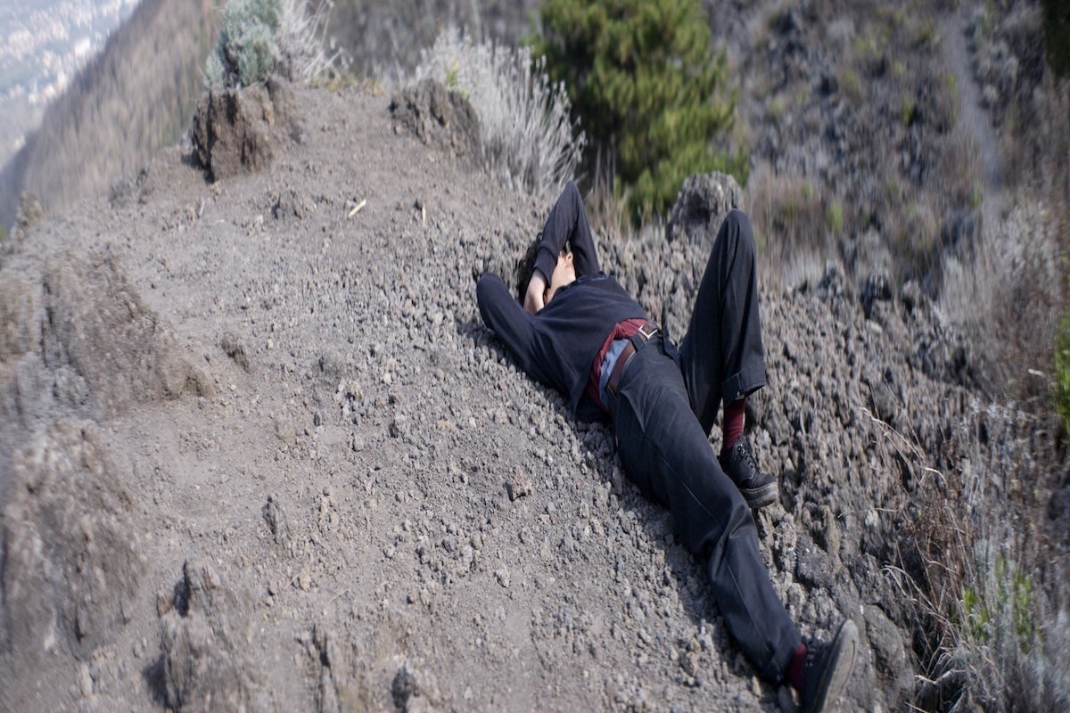 still of a woman dressed in all black lying on the slopes of Mount Vesuvius. Her right hand is behind her hand is behind her head and she raises her left arm over her head.