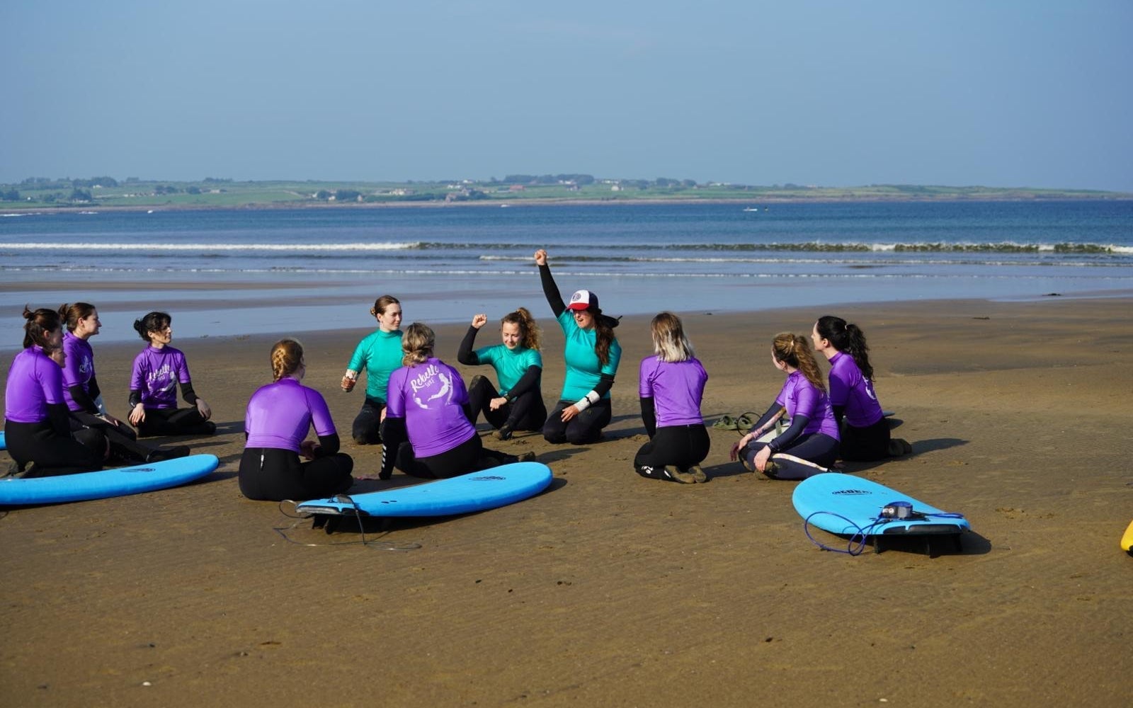 Group of surf students with surfboards sitting in a circle on the beach during a lesson