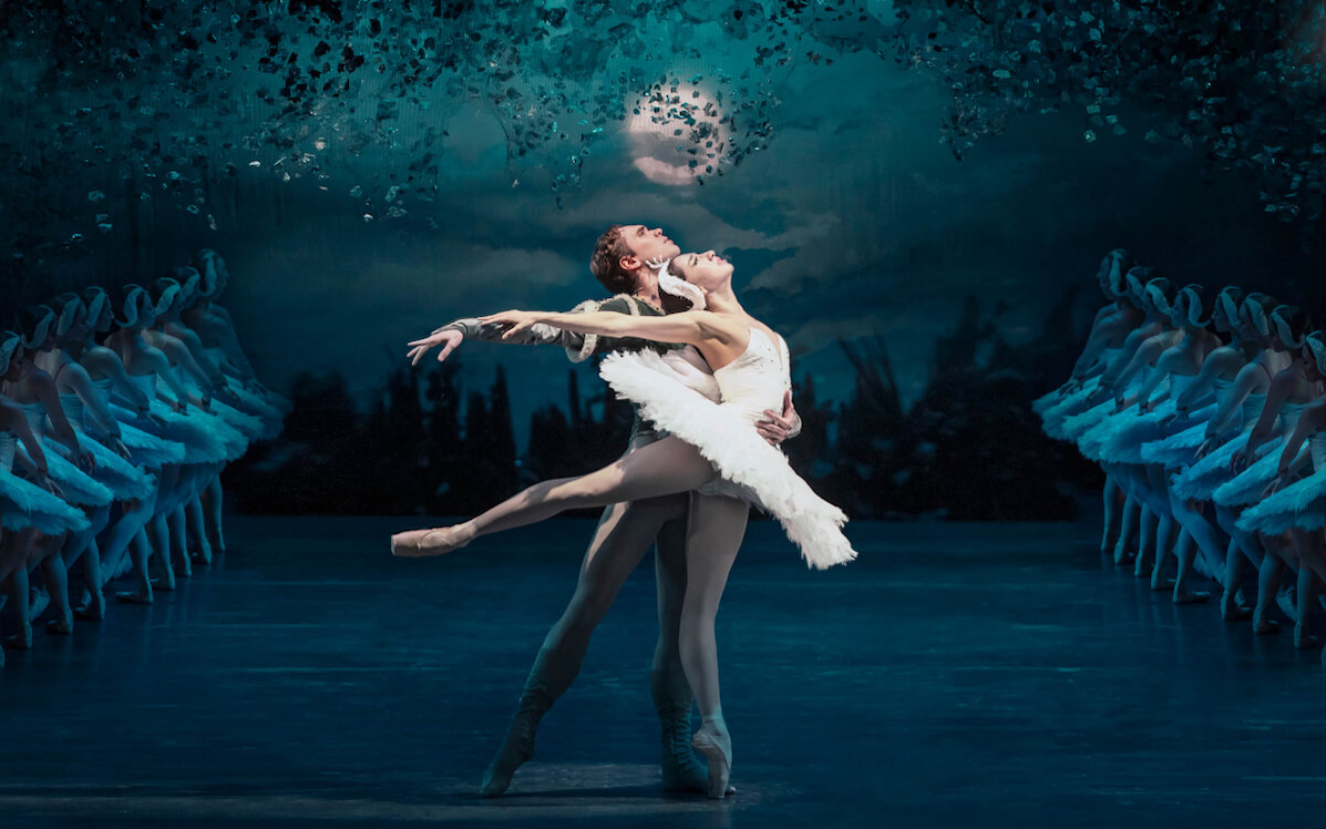 A female in short, white tutu on point ballet shows being supported by a man beside her on a dark stage