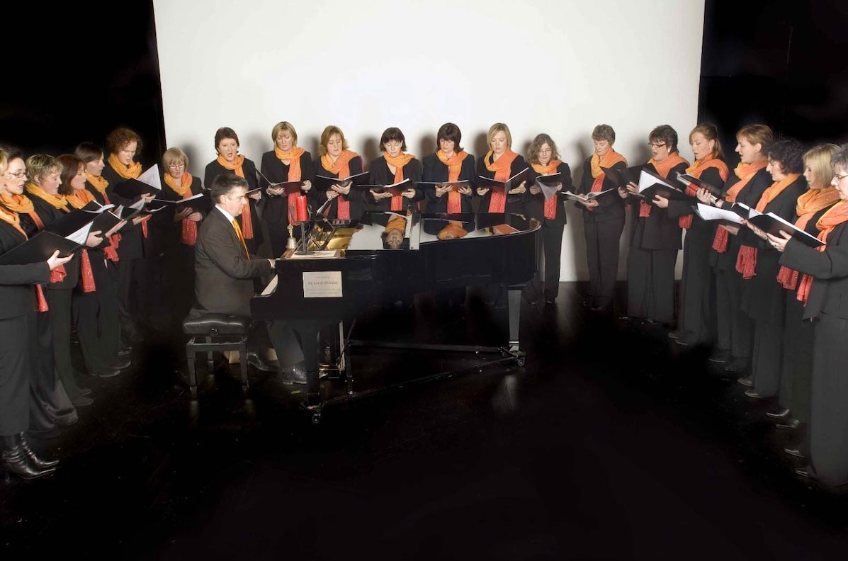 Festival of Voice: Cór Ban Cúil Aodha, a group of women standing in a semi circle around a small grand piano being played by a man.