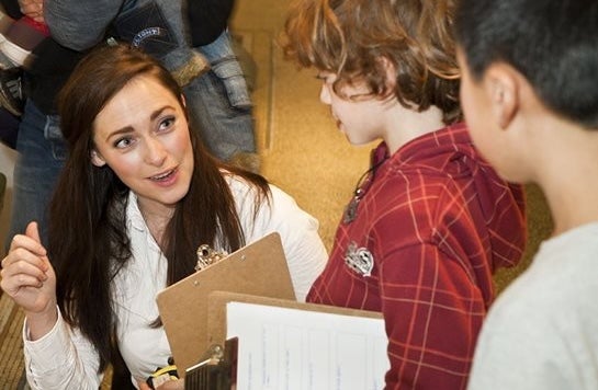 A woman with a clipboard is talking to 2 young children