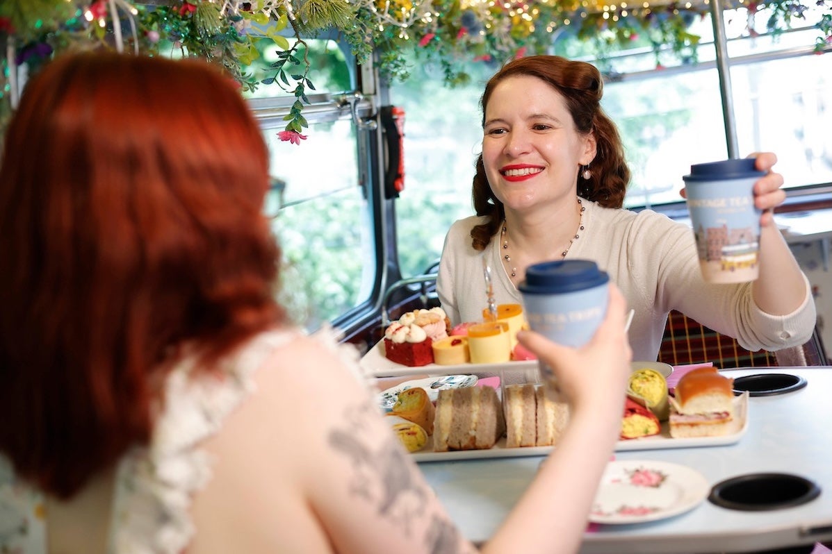 A woman seated at a table with mug and platter of sandwiches on top deck of double decker bus.