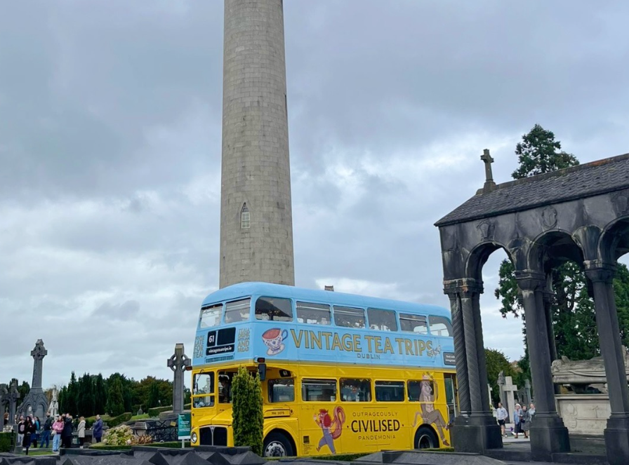 Vintage Tea Trips Bus at Glasnevin Cemetery, an old fashioned double decker bus park in a cemetery with tall thin round tower.