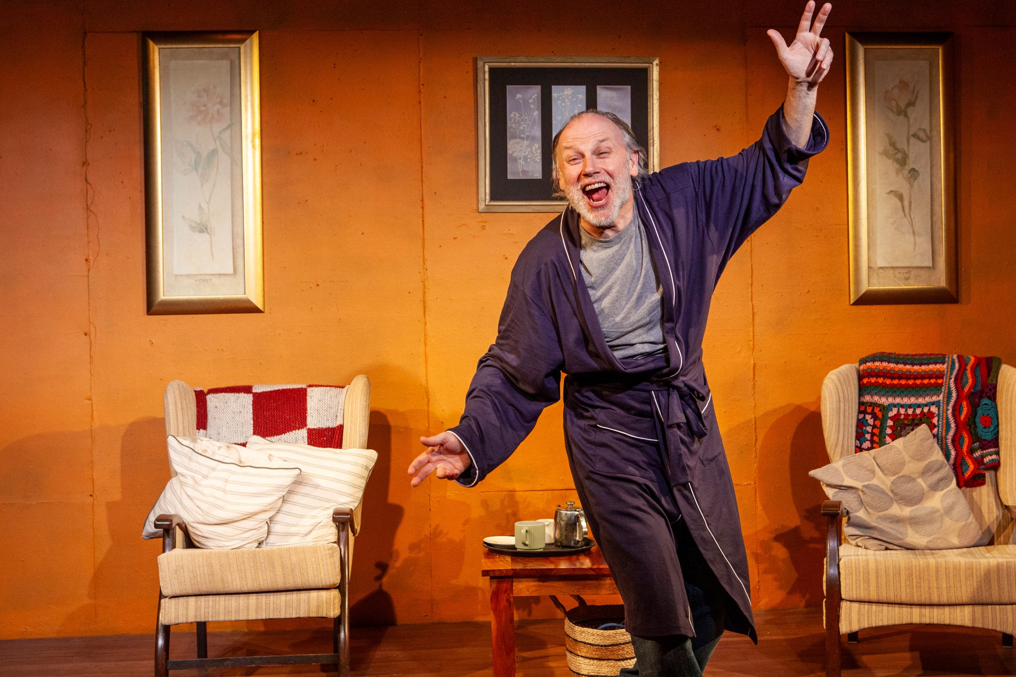 On a stage with armchairs, a man in a dressing gown is waving his arms looking excited.