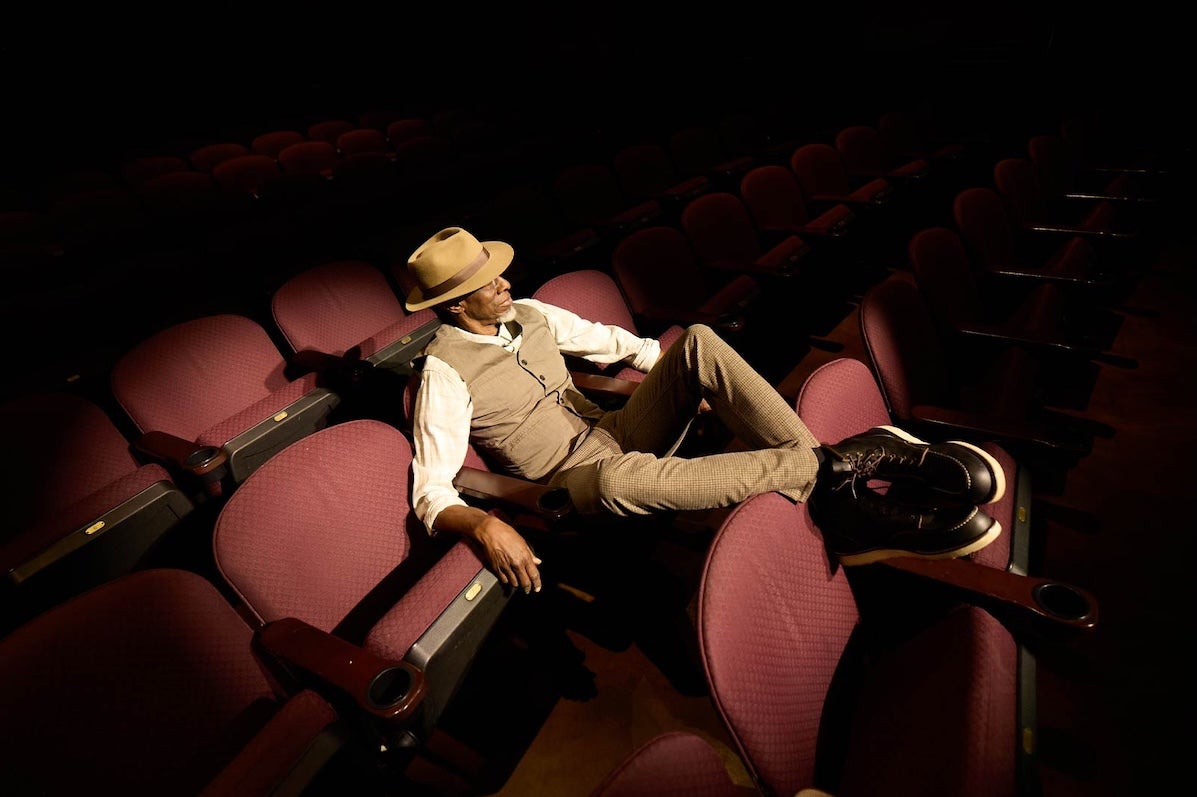 A man in waistcoat and shirt is seated in red cinema type seats with his feet up on the back on the seat in front, in darkness illuminated by a spot light.