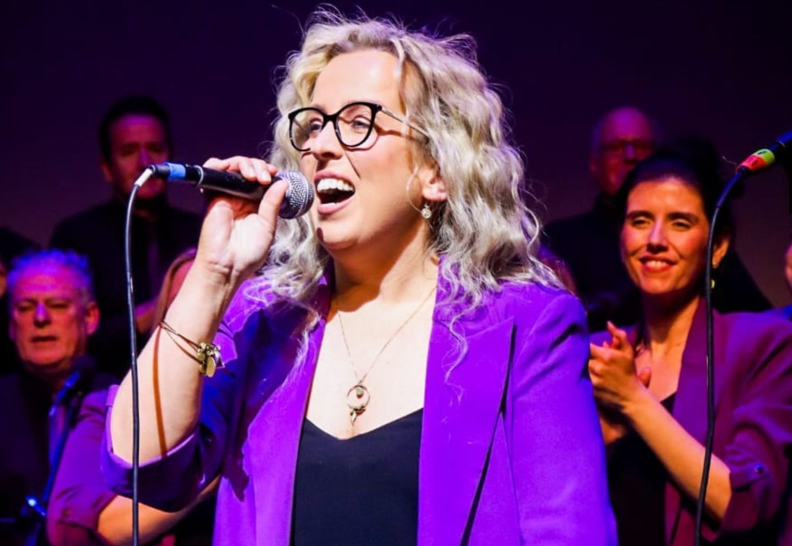 A woman in purple jacket singing with a hand held mic, with audience behind.