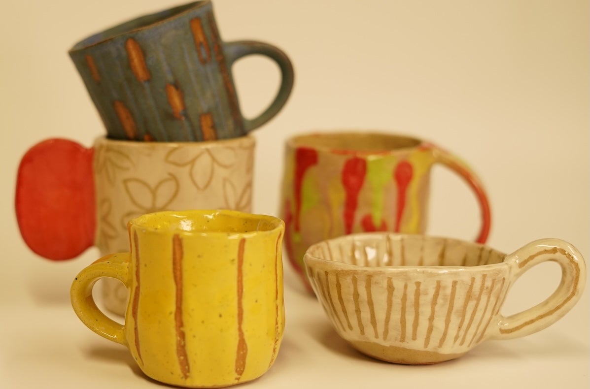 A group of 5 small, rustic handmade mugs in shades of yellow, beige, red and dark green.