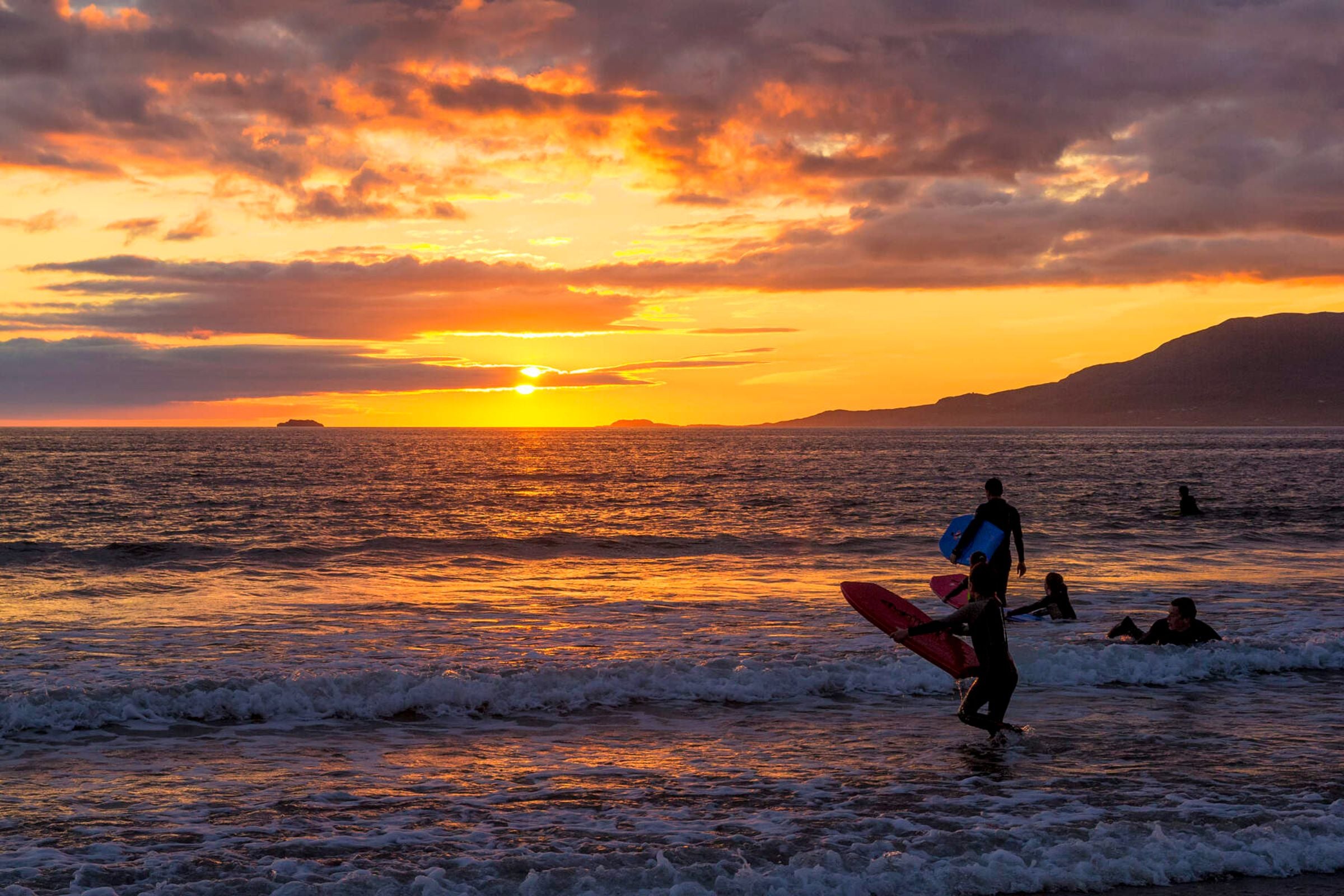 People running into the water at Carrowniskey Strand with surfboards at sunset.