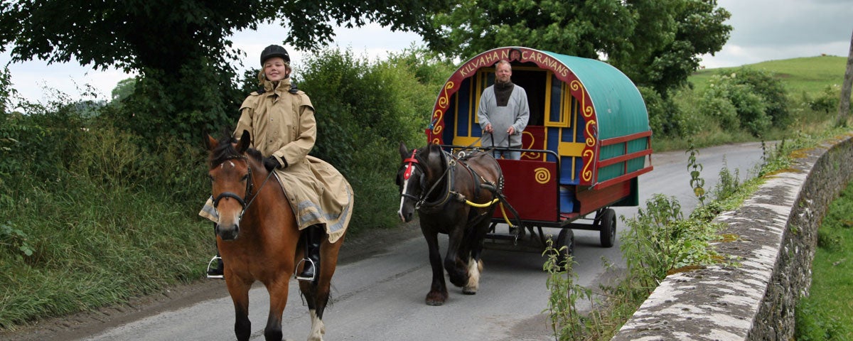 A horse drawn carriage riding along on the road with a woman on another horse riding in front