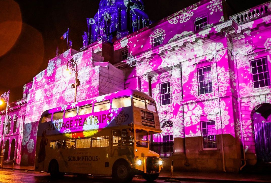At night a vintage double decker bus is parking outside a large out building the front of which is covered in pink lights with white snowflakes.