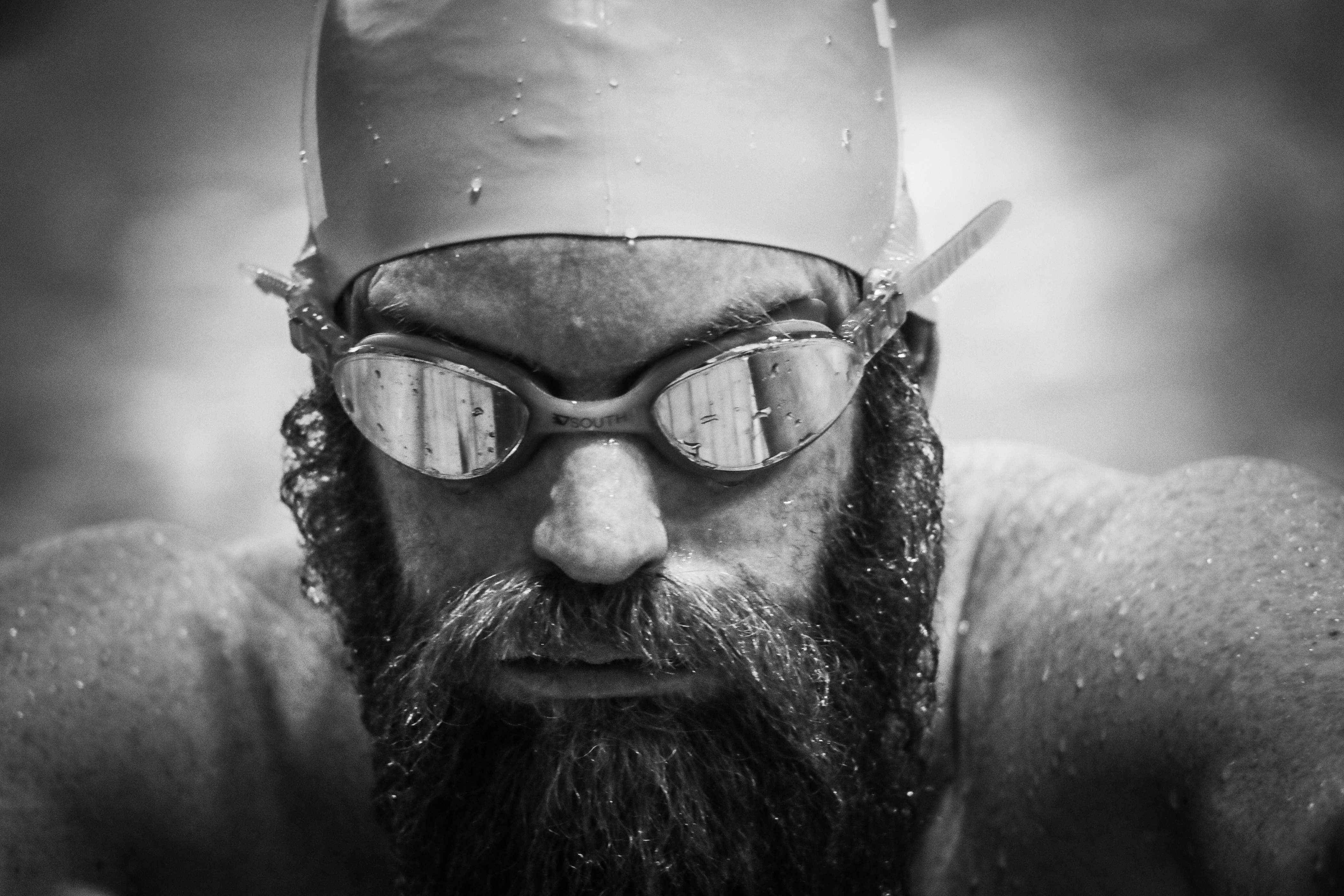 Close up headshot of man with a beard wearing swimming goggles and hat, covered in water droplets.