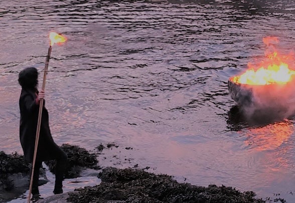 A person in a cloak holds a lit torch, watching a pire blaze in the middle of a misty river.