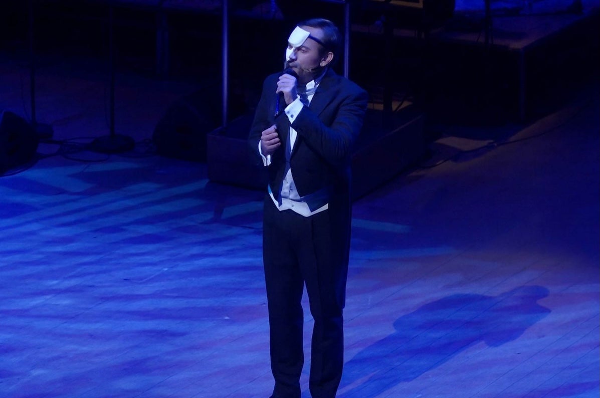 A man in evening dress singing into a mic on a dark stage, half of his face covered with a small mask.