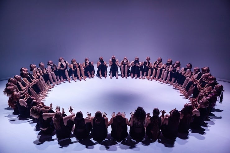 A large group of dancers seated on a white floor in a large circle.