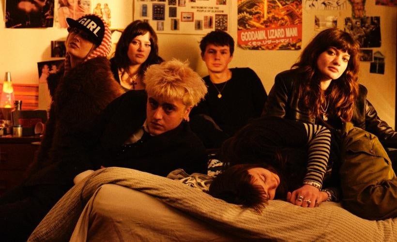 A group of 6 people seated on a bed in a room with poster on the wall behind
