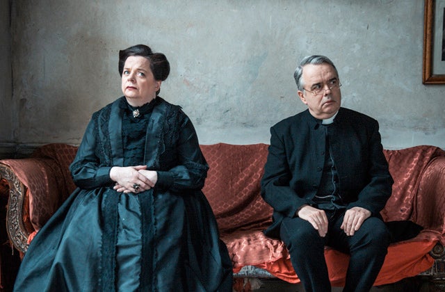 A serious looking man and woman dressed in black seated on a large sofa
