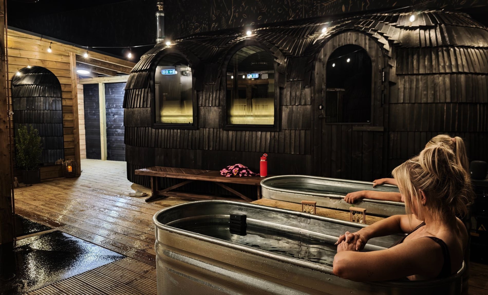 Two people sitting in cold plunge baths with mobile saunas to the right