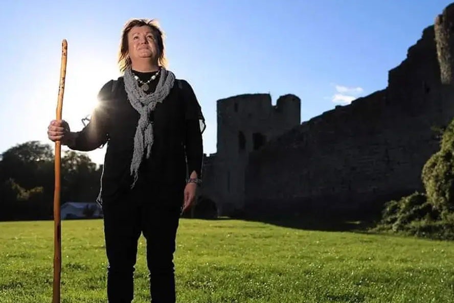A person standing in a field holding a tall stick against pale blue sky with outline of turret and walls.