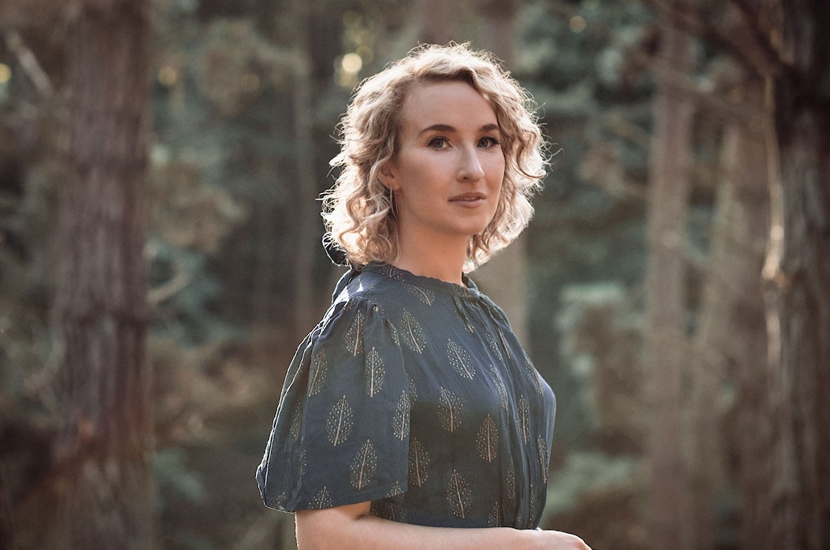 Slightly side profile on woman in dark, patterned dress with blurry trees in background.