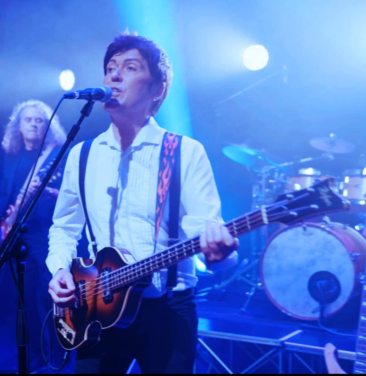 A man playing a guitar singing into a mic on a stand with band members behind