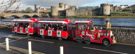 A small train on the road with a castle and the sea in the background