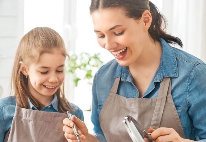 A woman and young girl wearing identical aprons are smiling, looking down at steaming saucepan on a stove