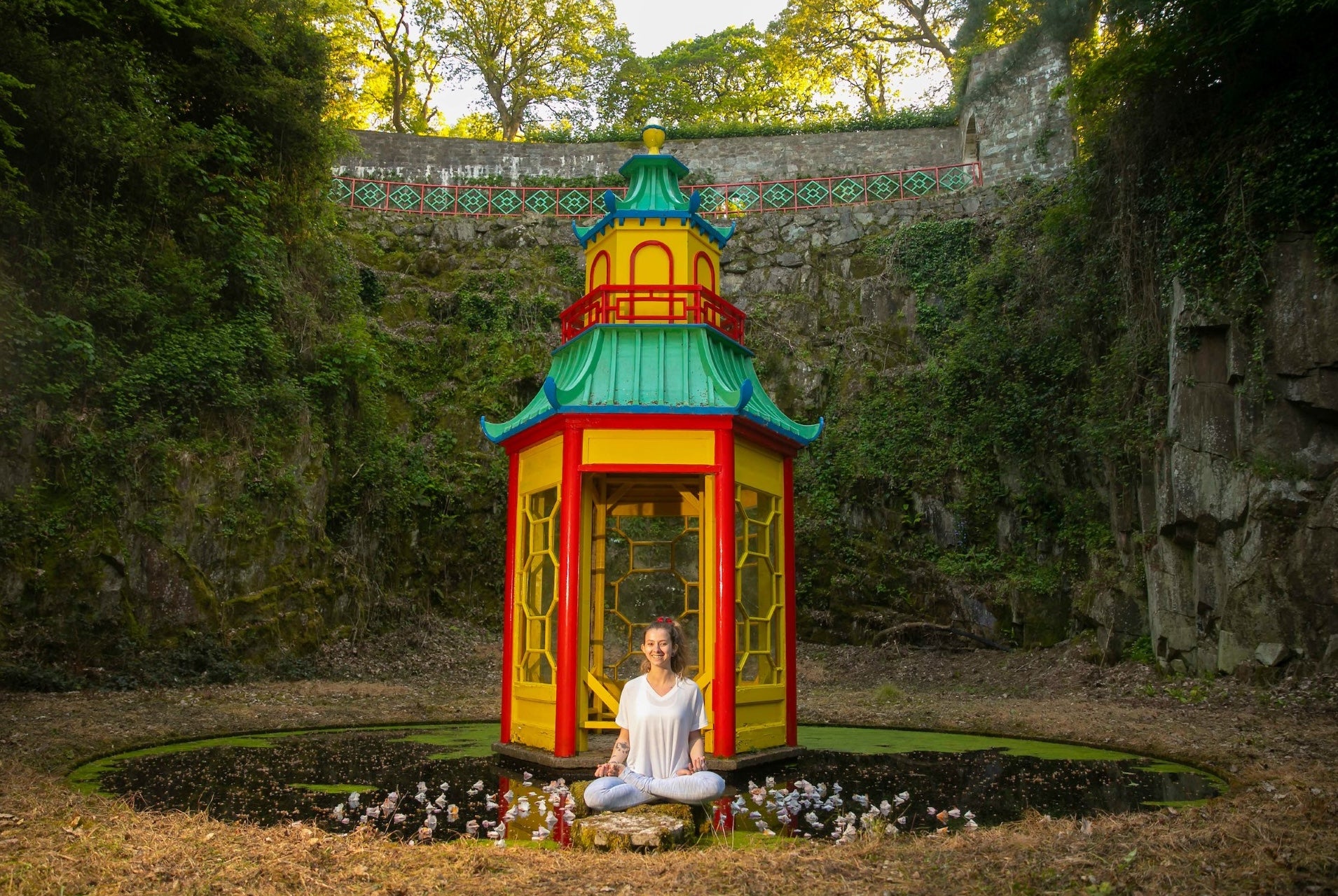 A smiling woman seated crosslegged in front of a small colourful pagoda on a small pond with high green covered walls all around.