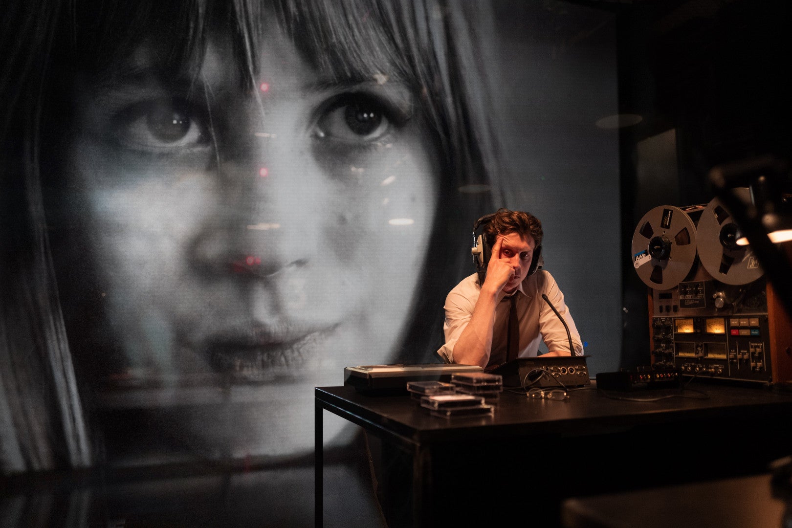 A man at a desk with audio equipment. An image of Marianne Faithful is being projected behind him.