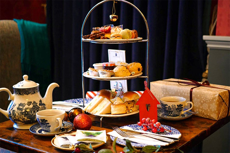 Festive Afternoon Tea stand displayed on a table in the Old Music Shop Restaurant, featuring scones, sandwiches, brioche buns, mince pies, miniature desserts, cups of tea, and a gift box, surrounded by Christmas decorations and ornaments.