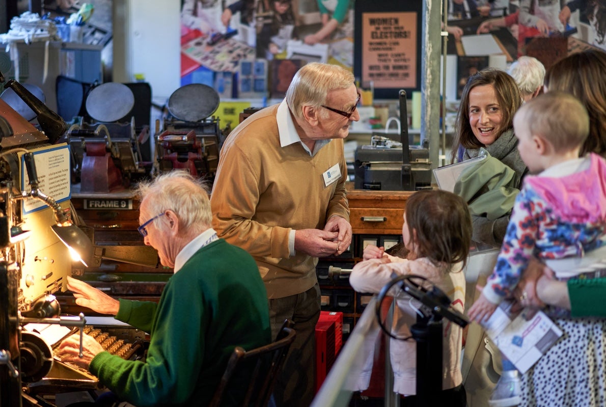 2 older men talking and demonstration old equipment to a small group of adults and children.