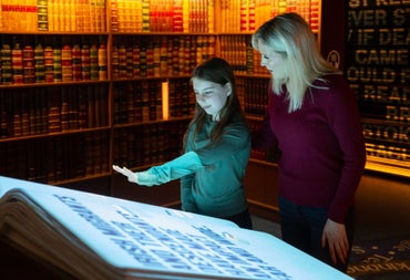 Two people standing looking at a large book in a room with lots of books on shelves