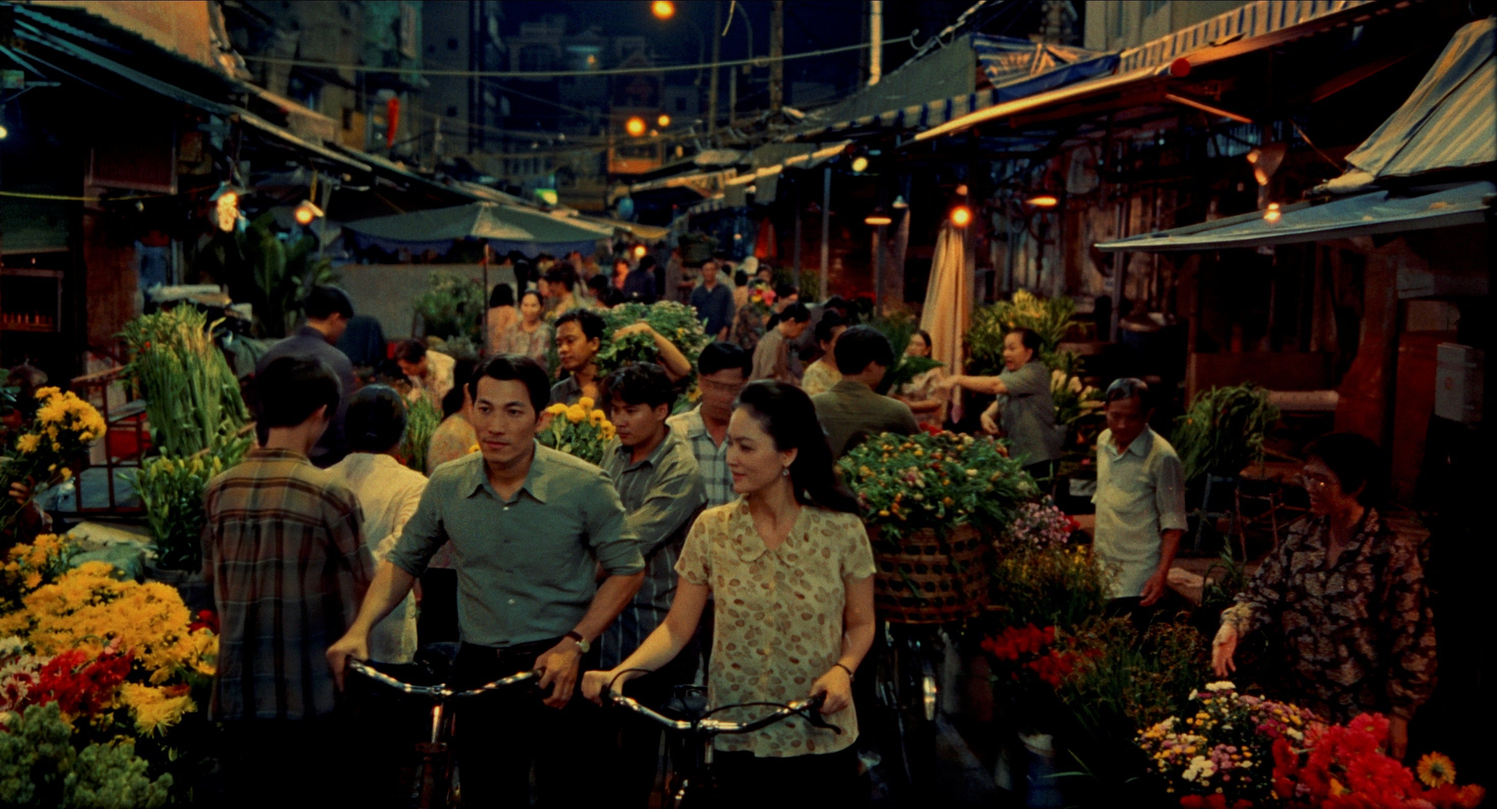 A man and a woman with bicycles are walking through a market.