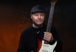 Christopher Cross, a man in a flat cap is holding upright an electric guitar.