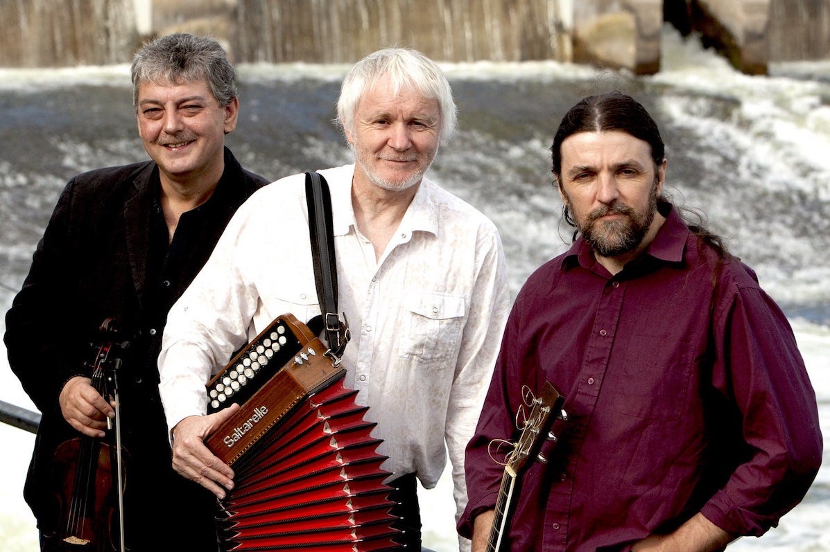Music Network presents Máirtín O’ Connor Trio, 3 men holding musical instruments standing against railings with cascading water behind them.