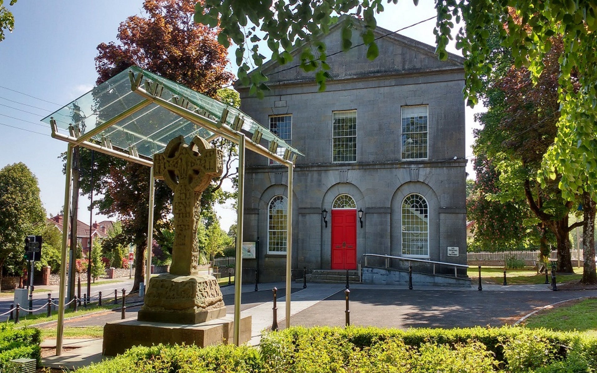 A two storey grey stone building with a red door and a Celtic cross in front