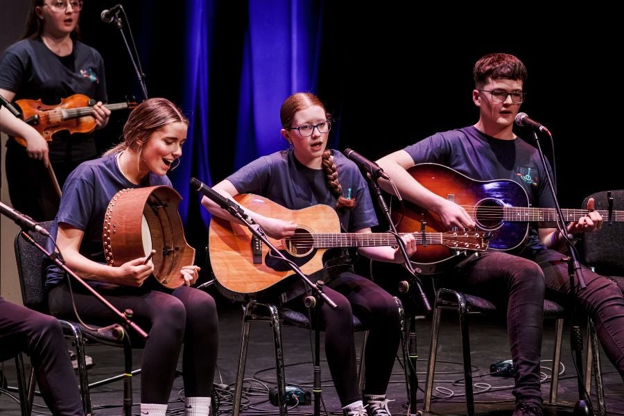 Young adults seated, playing musical instruments, singing as part of a larger group on a stage