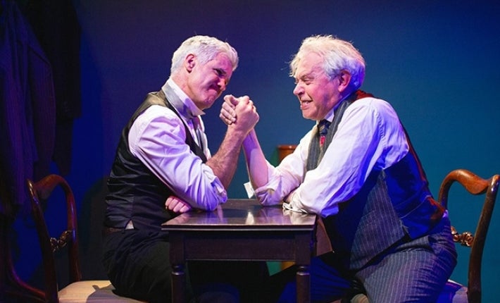 2 men in shirts and ties are sitting opposite each other, arm wrestling with gritted teeth.