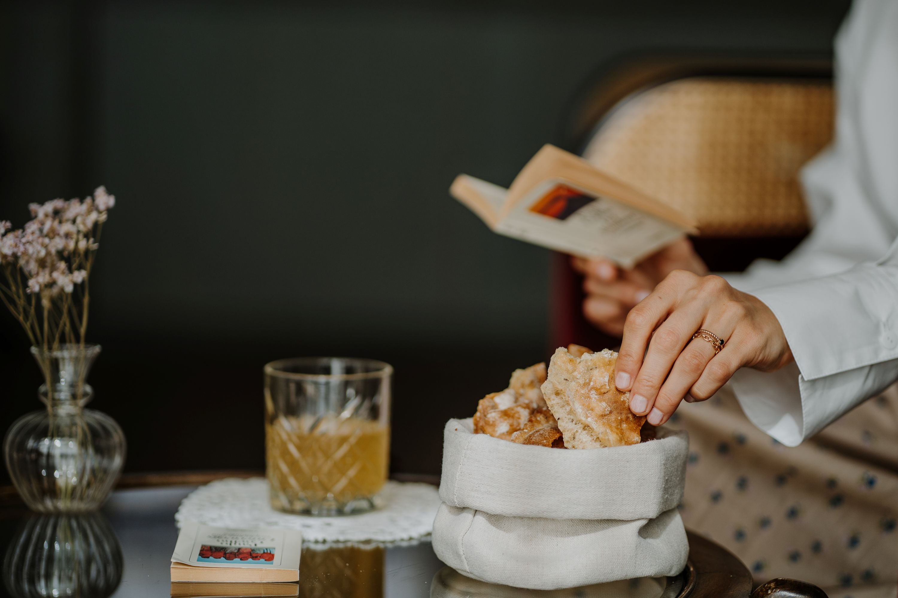 Person greift nach einem Schüttelbrot aus einem Körbchen auf einem Tisch mit Glas, offenem Buch und Blume in einer Vase.