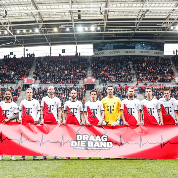 Voetballers van FC Utrecht en artsen van het UMC Utrecht staan voor een banner waarop 'Draag die Band' staat.