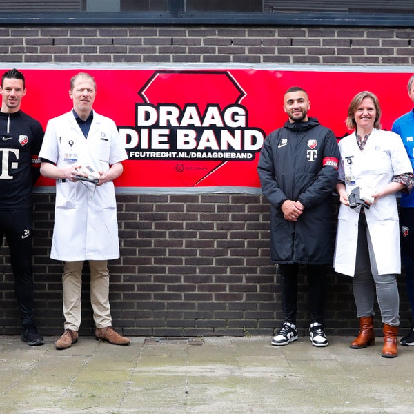 Voetballers van FC Utrecht en artsen van het UMC Utrecht staan voor een banner waarop 'Draag die Band' staat.