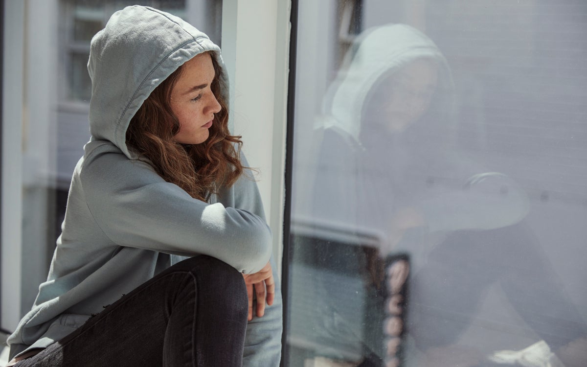 Een jonge vrouw in een grijze hoodie kijkt uit het raam.